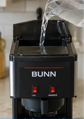 Fresh clear water being poured into the open top reservoir of a black BUNN-style coffee maker