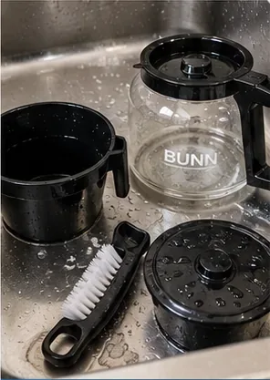 Coffee carafe, brew basket components, soft cleaning brush, and removable coffee-maker parts being washed in a sink