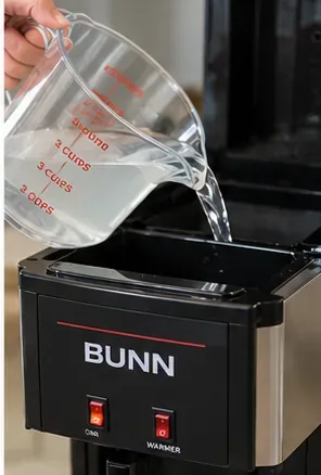 Clear liquid being poured from a measuring pitcher into the open top reservoir of a black BUNN-style coffee maker