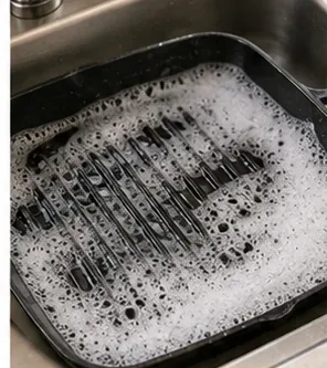 A black ridged grill pan sitting in a sink with warm soapy water loosening cooked-on residue.
