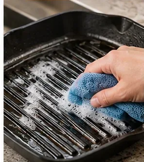 A hand using a soft cloth to scrub along the raised ridges of a black grill pan.