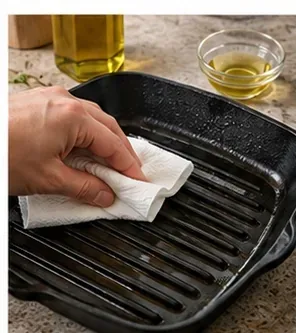 A very thin coat of cooking oil being wiped over a clean seasoned cast-iron grill pan with a paper towel.