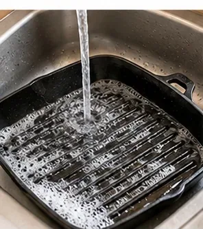A ridged grill pan being rinsed thoroughly under clean running water in a kitchen sink.