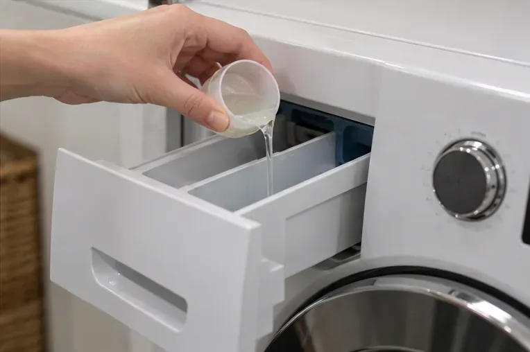 A small measured amount of clear liquid detergent being poured into a washing machine detergent drawer