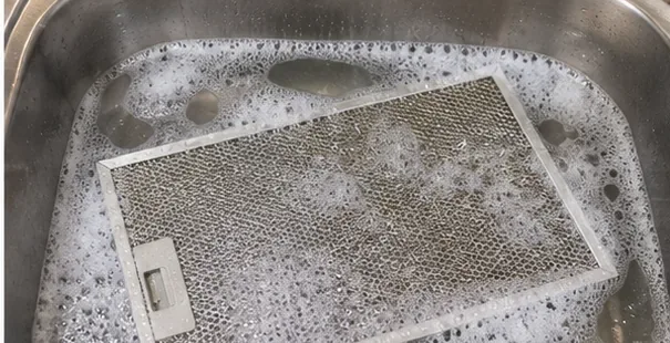A rectangular metal range hood grease filter soaking flat in a sink filled with hot soapy water
