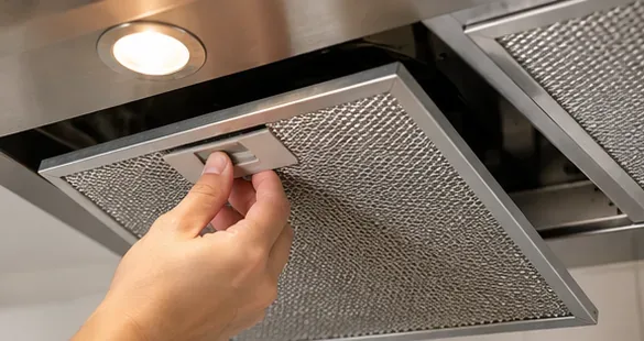 A hand releasing a rectangular metal mesh grease filter from the underside of a stainless steel range hood