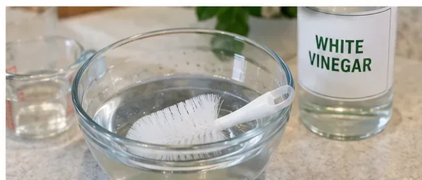 A bowl, soft cleaning brush, measuring cup, and bottle labeled white vinegar on a kitchen counter.