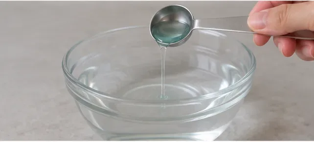 A small measured amount of liquid detergent being poured from a metal measuring spoon into a clear bowl of water.