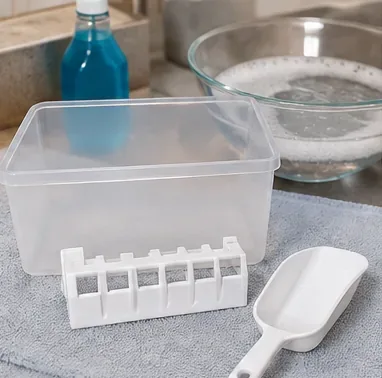 Un recipient pentru gheață curat și transparent și piese detașabile din plastic ale aparatului de gheață așezate pe un prosop lângă un bol cu ​​apă cu săpun