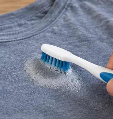 Periuță de dinți moale care freacă ușor o cantitate mică de detergent spumos în reziduurile de autocolant de pe o cămașă gri-albăstruie