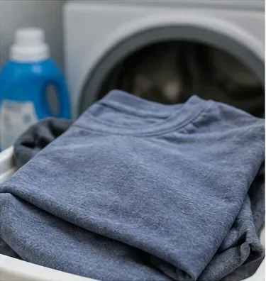 Cămașă gri-albastră împăturită lângă o mașină de spălat cu încărcare frontală deschisă și o sticlă de detergent de rufe