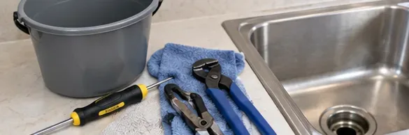 Bucket, pliers, screwdriver, towel, and basic tools arranged beside a stainless steel kitchen sink before drain removal