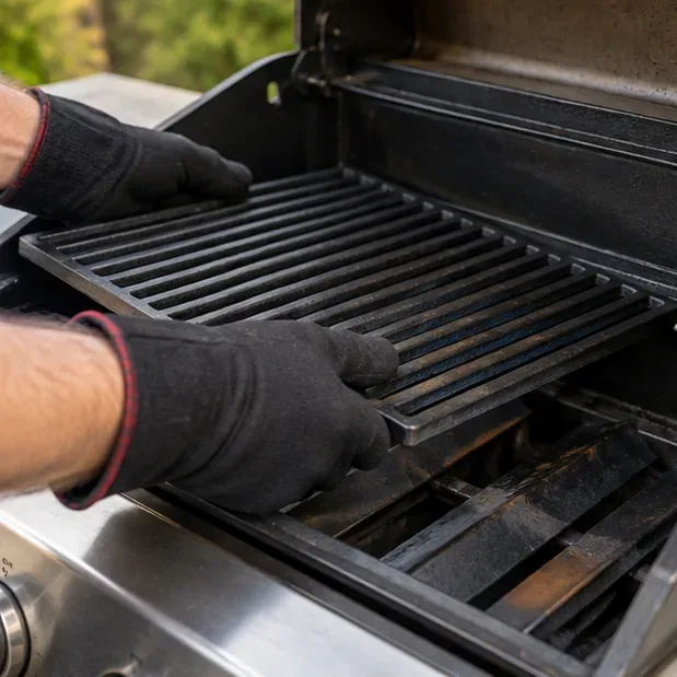 Gloved hands place the cleaned and lightly oiled cast iron grate back into the open gas grill before the heating cycle.