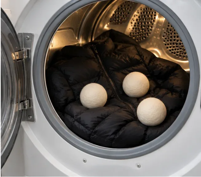A black puffer sits in a tumble dryer with three clean wool dryer balls for low-heat drying.
