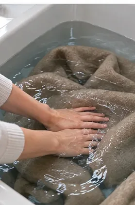 Hands rinsing a submerged wool coat in clean water inside a sink.