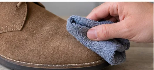 Microfiber cloth gently wiping a small area on the toe of a tan suede shoe.